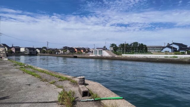 Scenic wide view of a calm waterway and concrete wharf with coastal townscape under a cloudy blue sky in Kurobe Port, Toyama, Japan. Ideal for regional infrastructure and web backgrounds.