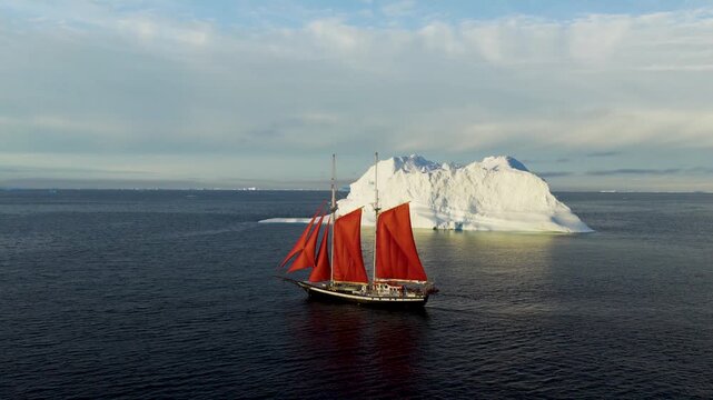 Aerial view of a sailboat with striking red sails navigating the cold arctic waters near a massive white iceberg under a cloudy sky in Greenland.