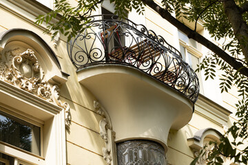 Ornate wrought iron balcony with curved railing above sculptural stone entrance on cream classical building framed by leafy branches, decorative European architecture facade © Vlad Yakubovskiy