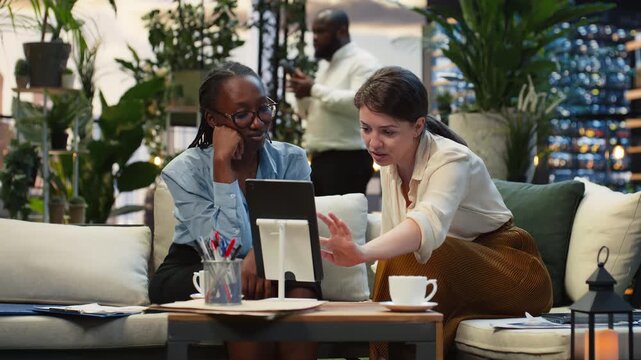 Company coworkers look at a tablet screen in a stylish rooftop workspace while reviewing work notes; businesswomen compare project information on the device above the business district.