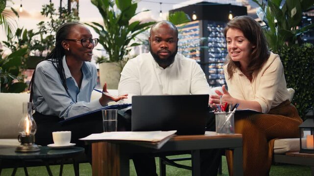 Happy business colleagues smile while reading good news on a laptop in a rooftop lounge; cheerful professionals are pleased by positive company performance metrics during project work.