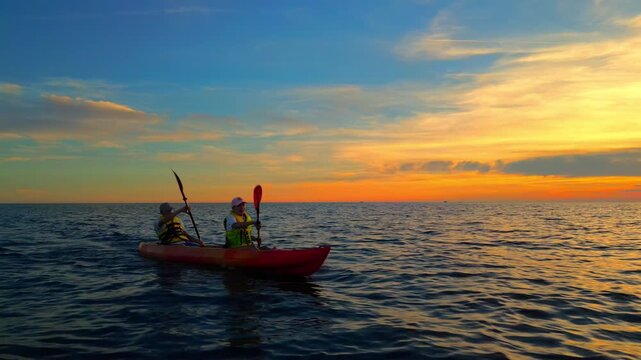 Two kayakers, a couple, paddle through the calm sea at sunset, embracing an adventure in a serene, golden setting. Their synchronized strokes and shared smiles highlight an active lifestyle.