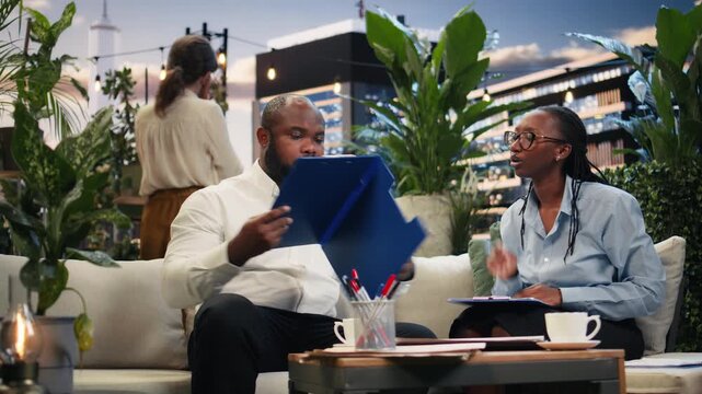 Happy businesspeople discuss project paperwork and sign a contract in a rooftop lounge, then shake hands after completing the business deal agreement.
