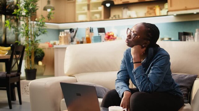 Pensive woman works from home on laptop while uncertain about tasks, frowning at screen during ambiguous teleworking and multitasking in living room.