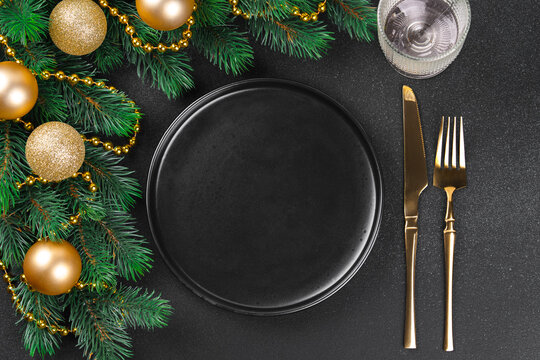 Christmas table setting with empty black plate, golden cutlery, green spruce branch with gold balls, on dark background. Template, mockup with festive aesthetic holiday atmosphere.