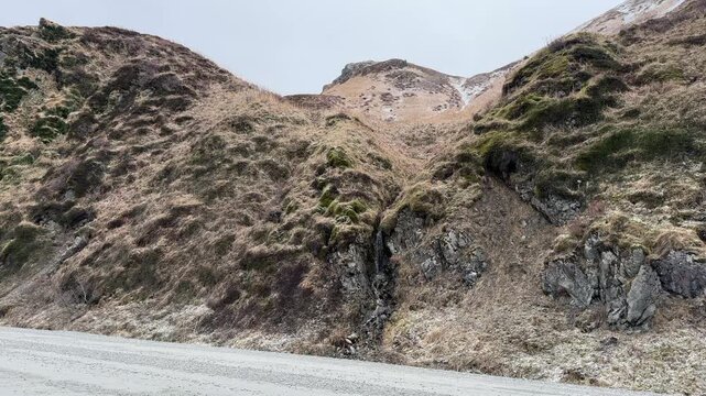 Low angle shot of steep rocky cliffs covered in moss and tundra with a small waterfall flowing next to a gravel pass road.