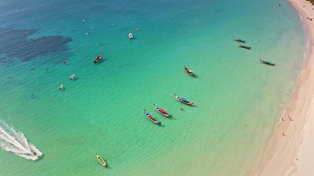 Aerial view shot of tropical beach in Phuket, Thailand. People enjoying clear turquoise water, longtail boats anchored in bay and a jet ski creating wake near white sand shore. Summer travel and relax