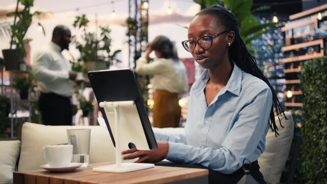 Close-up shows a businesswoman using a tablet on a stand at a rooftop workspace table while coworkers talk in the background, as she reviews digital project information during a work session.