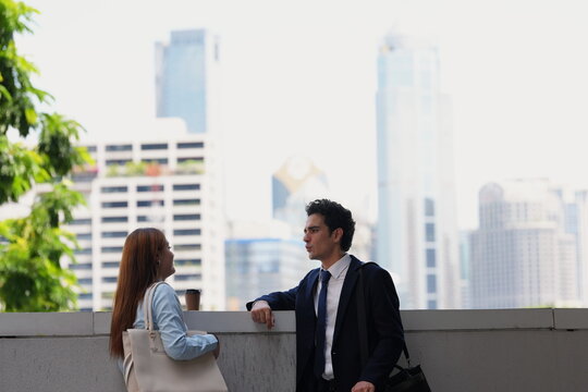 Two young professionals take a coffee break at a parapet overlooking the city, a strong skyline image for real estate, careers and lifestyle campaigns, with hazy towers behind