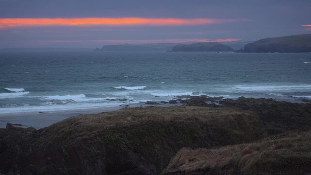Rolling waves approach a rugged rocky shoreline beneath a narrow band of orange sunset light in Pembrokeshire.