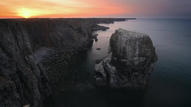 A massive offshore rock formation rises beside steep limestone cliffs beneath a glowing sunset sky in Pembrokeshire.