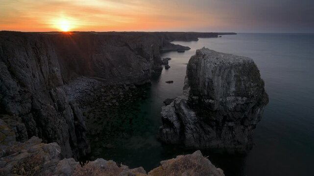 Timelapse of changing sunset light above a towering sea stack, steep coastal cliffs and calm water in Pembrokeshire.