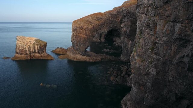 A detached limestone sea stack stands beside towering coastal cliffs surrounded by calm blue seawater in Wales.