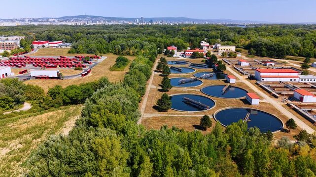 Row of circular wastewater basins beside a service road. Treatment pools recede toward buildings, with trees in the foreground and an urban horizon beyond.