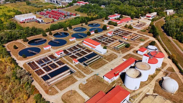 Wastewater treatment basins and tanks viewed from above. Dark circular pools and open channels form parallel rows beside white structures with red roofs.