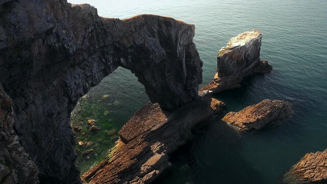 A dramatic limestone arch and isolated sea stack rise above dark blue water on the Pembrokeshire coast.