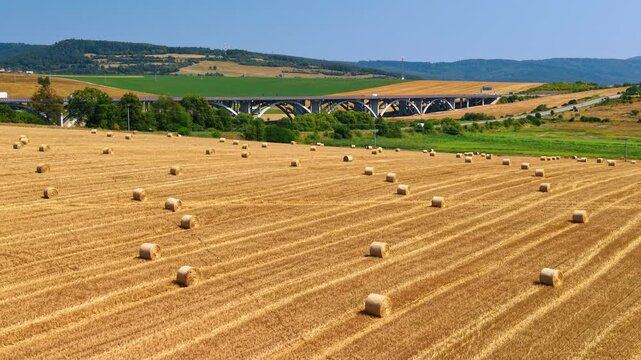 Straw bales in a field below an arched road bridge. Round bundles lie on golden stubble, with trees, concrete spans and patchwork hills in the background.