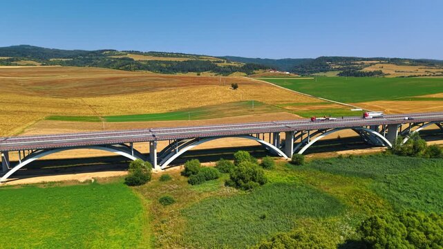 Arched motorway bridge crossing agricultural fields. Vehicles travel over concrete supports above green crops, with harvested land and low hills beyond.