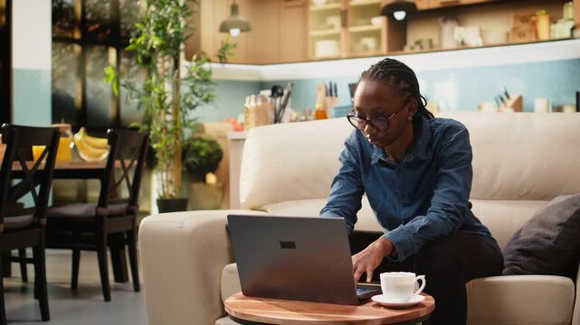 Black woman works remotely from home setup while drinking coffee and solving daily administrative tasks on laptop and internet. The digital nomad follows a flexible work routine at home.