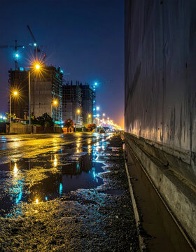 Night city street with construction site, wet pavement, and colorful light reflection beside concrete wall
