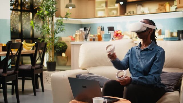 Woman freelancer uses VR headset for metaverse immersion and motion tracking during multitasking. Person telecommutes with virtual reality at home, combining domestic life with laptop-based work.