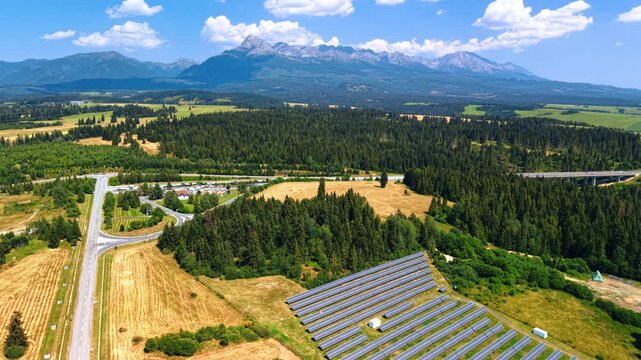Solar panel rows beneath a mountain range and forest. Reflective modules stand beside a road junction and golden fields below Krivan in the High Tatras.