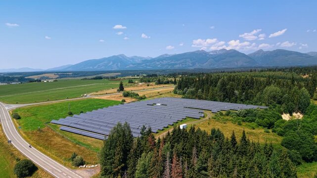 Solar farm between a curving road and conifer woodland. An aerial view shows parallel modules across a grass plot, with fields and a mountain ridge behind.