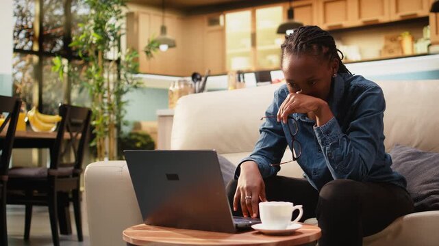 Black woman with headache works from home feeling overwhelmed by tasks and responsibility, drained by discomfort and eye strain. Adult rubs her temples indoors while showing visible physical fatigue.