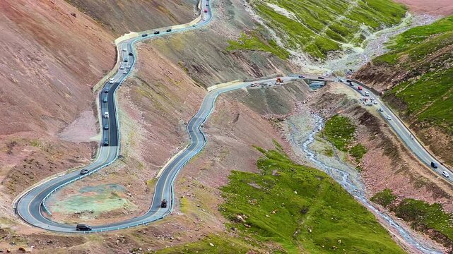 High angle shot shows a winding mountain highway with cars traveling along steep, rocky slopes and grassy valleys under bright day.