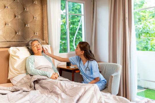 Asian female doctor examine senior woman patient with ear infrared thermometer measure temperature. Caregiver or nurse visit elderly patient health at home. Home medical healthcare service concept.