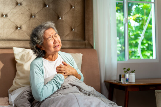 Asian senior woman suffering chest pain or heart attack on bed. Elderly patient holding chest with painful expression, heart problem, health emergency, cardiovascular disease and medical care