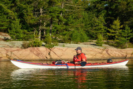 Sea kayaker pauses in a red kayak along a pink granite shoreline below pine forest in morning light on Georgian Bay, Ontario, Canada