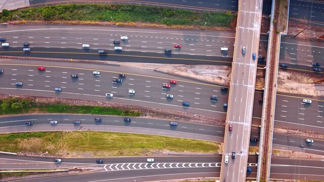 The geometric lines of a complex highway junction where multiple roadways run alongside each other. Cars navigate the distinct asphalt lanes under a long overpass bridge and a parallel train track.