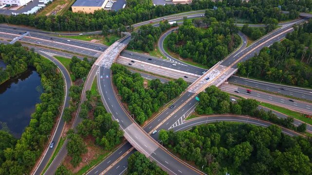 This bird's-eye on a large, winding cloverleaf interchange surrounded by a dense blanket of lush green trees. Cars travel by the roads across green lush forests.