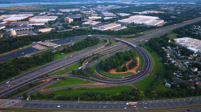 A series of massive industrial warehouses and commercial parks built right alongside a looping highway system. In the far distance, a wide river or bay cuts across the horizon under a hazy sky.