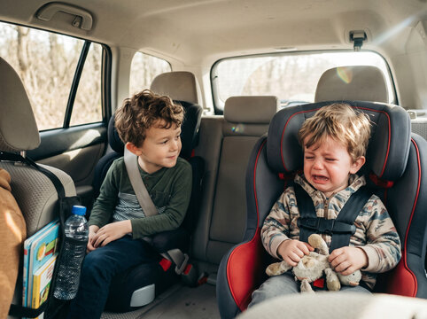 Two young brothers sitting in car seats in the back of an automobile during a family road trip, with the older boy looking at his crying toddler brother who is upset and holding a stuffed toy animal