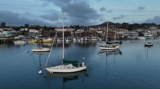 Morro Bay California sailboats anchored in the marina harbor with waterfront town buildings and overcast sky 4k aerial drone video