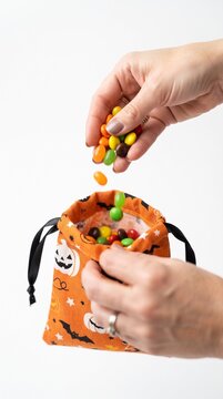 Hands filling a Halloween drawstring bag with colorful candy. The bag has a festive orange background with bat and pumpkin motifs