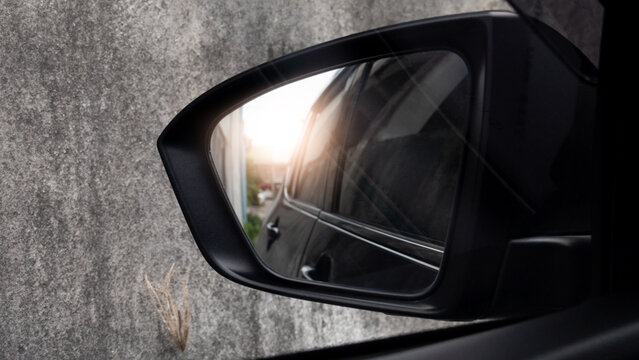 Close up side view of a modern black car side mirror reflecting an outdoor urban environment and lighting. Side mirror is a concrete wall featuring a reflection of the village in the glass.