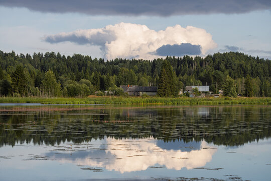 A summer rural landscape. View of wooden houses located on the shore of a lake (a rural reservoir). Clouds are reflected in the water. Oleshkovka village, Nyuksensky district, Vologda region, Russia.