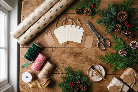 Christmas wrapping paper, gift tag, twine, scissors and evergreen branch on rustic wooden table.