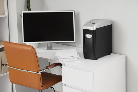 Modern shredder with documents and computer on desk in office interior