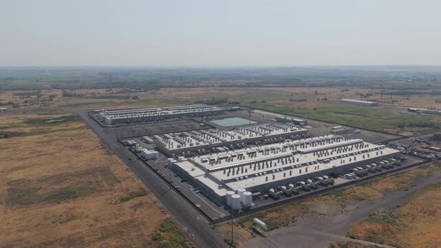 Aerial view of the Amazon Web Services data center with industrial buildings, cooling units, electrical substations, and arid landscape.