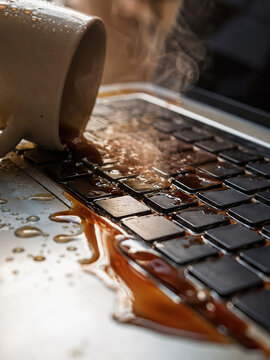 A mug of coffee spills onto a laptop keyboard, creating a spreading pool of dark brown liquid and visible steam.