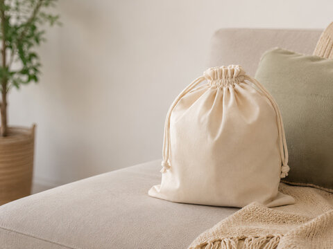 A beige drawstring bag rests casually on a light-colored sofa in a minimalist room The bag is made of a natural, off-white fabric and has a knotted drawstring closure.