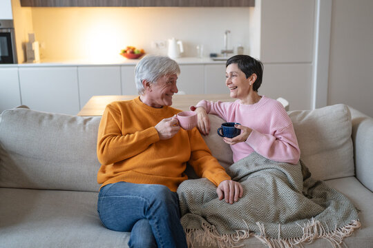 Happy elderly husband and wife sit close under warm blanket, drink tea and laugh together, affectionate senior couple enjoys conversation, comfort and shared leisure time at home in retirement.