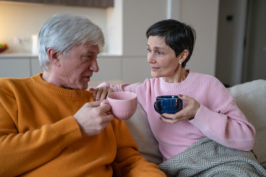 Senior husband and wife sit on sofa holding colorful cups, smiling elderly couple enjoys tea and friendly conversation, married partners spend relaxed time together and share daily life.