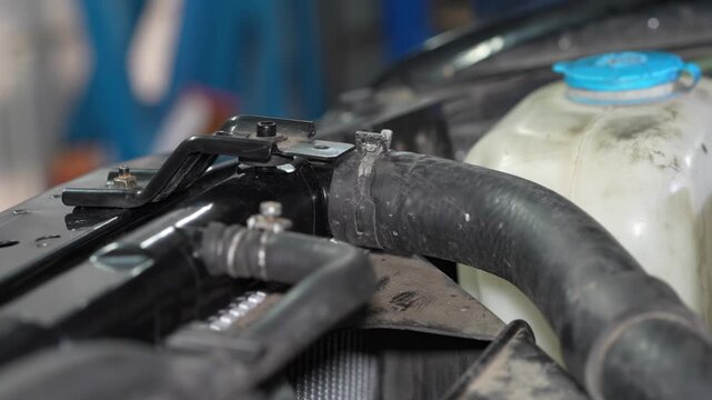 A close-up shot of a car radiator upper tank, showing the main coolant hose with a spring clamp and a smaller overflow hose connected near the expansion tank.