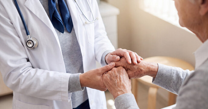 Compassionate doctor comforting elderly patient by gently holding wrinkled hands inside bright clinical consultation room