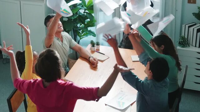 Top view of smart people throw paper and giving high five at meeting table with financial document and stock market analysis paper scattering around. Aerial view of team celebrate project. Symposium.